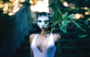 Image shows a women in a white mask behind green plants. Greenwashing. 